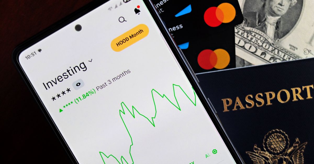 Smartphone displaying investing app, with credit cards, cash, and passport nearby, symbolizing finance